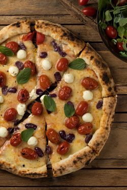 Pizza Mamma Quaresma sobre mesa de madeira. Leva queijo de cabra, tomate cereja, azeitonas pretas e filhas de manjericão.